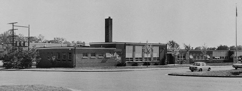 Black and white photograph of the front of Wakefield Forest Elementary School taken in 1965. The building looks relatively new and small trees have been planted all along the front. Two early 1960s era cars are parked in front of the building. 