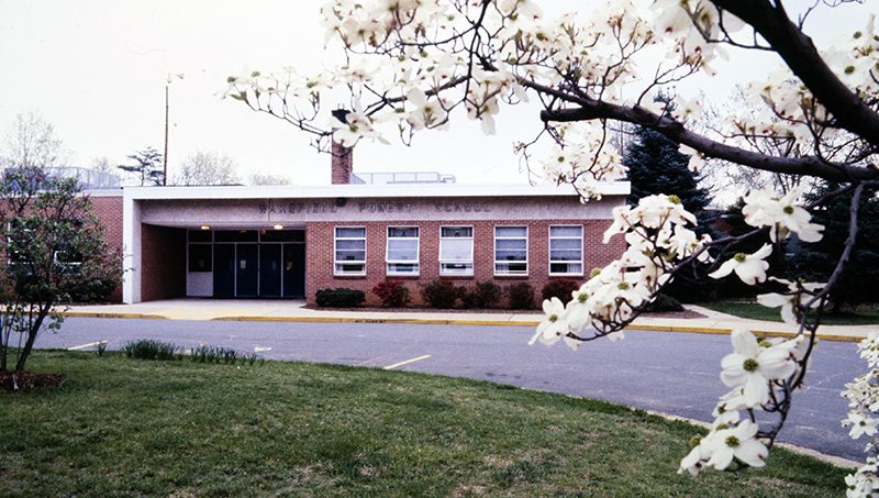 Color photograph from a 35 millimeter slide showing the main entrance to Wakefield Forest Elementary School. The picture was taken sometime during the late 1970s or early 1980s. It is springtime and the dogwood tree in front of the school is in bloom. 