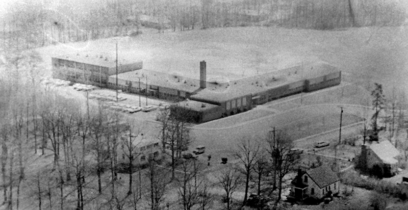 Black and white aerial photograph of Wakefield Forest Elementary School taken from the north. You can see old homes built near the school that have since been torn down. The building is shaped like the letter L. It is mostly one-story, but the far left portion of the building is two-stories tall. The main entrance to the building appears to face Virginia Avenue, not Iva Lane as it does today. 